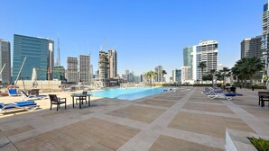Un área de piscina en la azotea con sillones y mesas, rodeada de rascacielos y palmeras.
