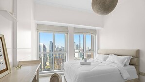 Un dormitorio con una ventana grande que ofrece vista a la ciudad, una cama con ropa blanca, un escritorio y una silla.