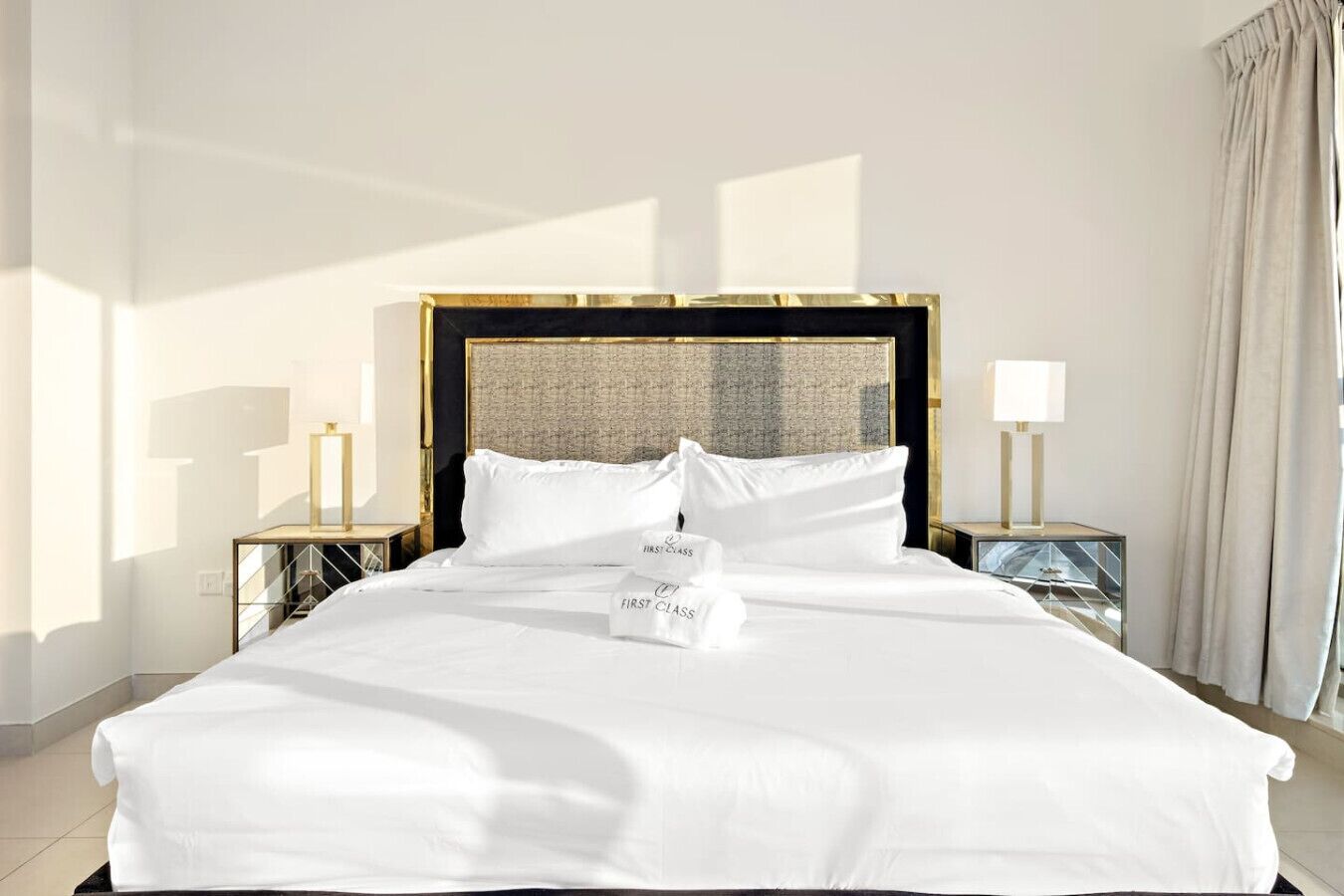 Een netjes opgemaakt bed met witte lakens en kussens, een gouden hoofdbord en twee nachtkastjes met lampen.