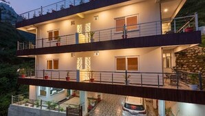 Un edificio de varios pisos con balcones, un coche estacionado adelante y un muro de piedra en un costado.