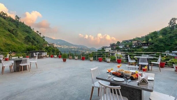 Un área de comedor al aire libre con mesas y sillas, rodeada de follaje y colinas al fondo.