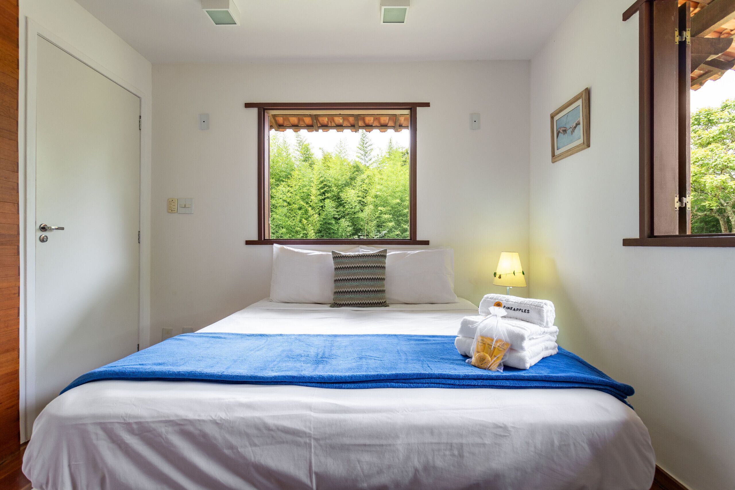 Ein Hotelzimmer mit Bett, Fenster mit Blick auf Bäume, einer Lampe und einem Handtuchhalter mit Handtüchern.