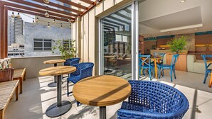 Un espace extérieur aménagé avec des tables en bois et des chaises en osier bleu, une pergola en bois ornée de guirlandes lumineuses, et une porte en verre donnant accès à un espace intérieur.