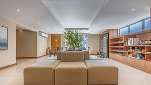 Un hall d’hôtel moderne avec un canapé modulable, une étagère en bois contenant des collations et une grande fenêtre laissant entrer la lumière naturelle.