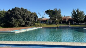 A rectangular swimming pool with clear blue water, surrounded by a paved deck and a grassy area with trees and buildings in the background. - Vilanova by Check-in Portugal (Albufeira)
