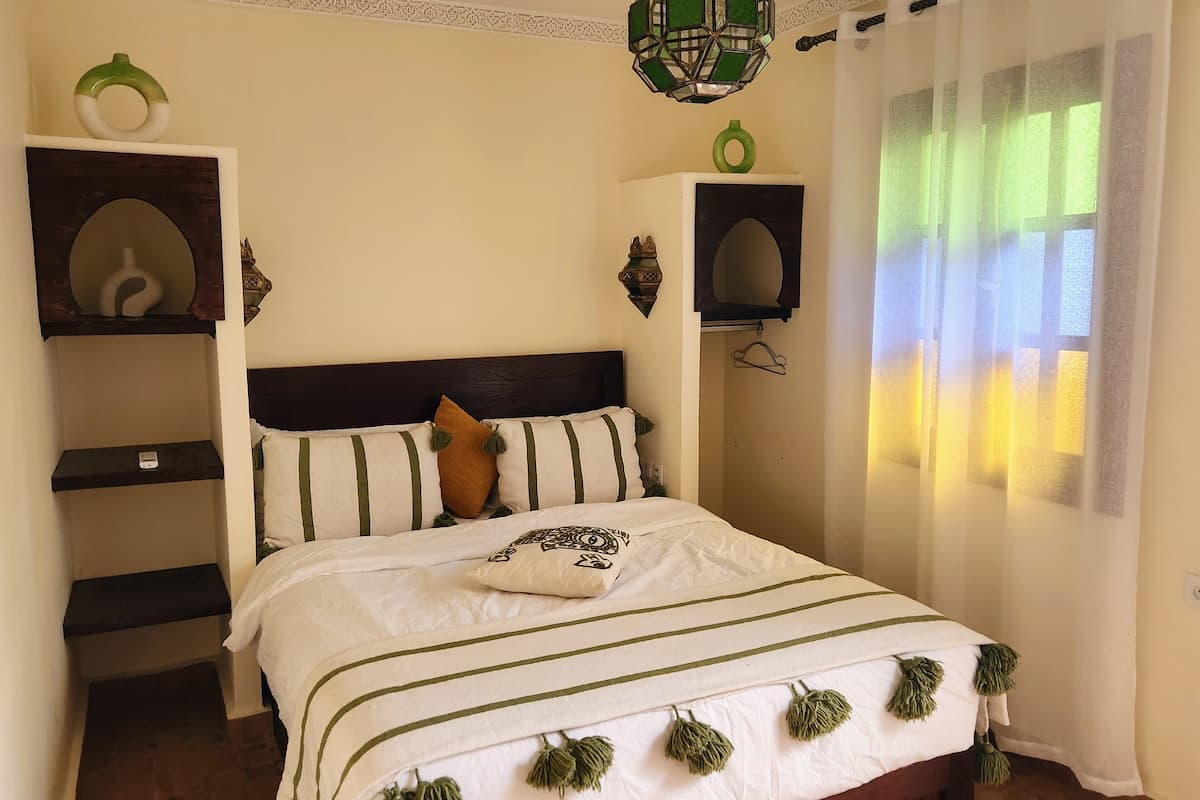 Una cama bien hecha con ropa de cama a rayas y flecos, un cabecero con soportes decorativos de pared, y una ventana con cortinas translúcidas que dejan entrar luz natural.