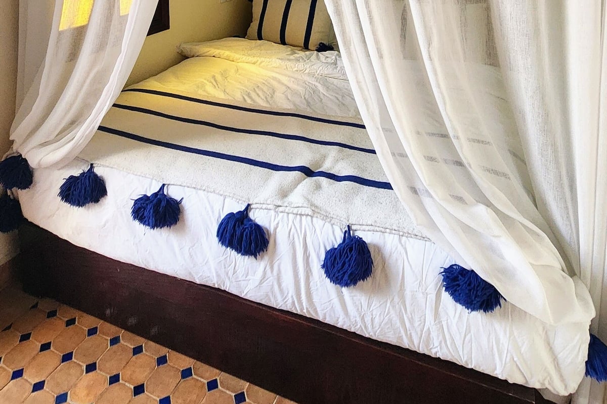 Un dormitorio con cama, cortinas blancas y suelo estampado.
