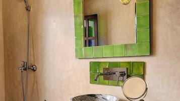 Un baño con pared de azulejos verdes, un lavamanos blanco con recipiente, un cabezal de ducha de mano, un espejo y dos toallas en un tendedero.