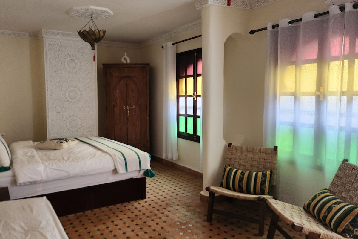 Un dormitorio con cama, una silla, un armario de madera, una ventana con vidrieras de colores y un suelo con estampado.