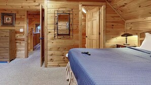 A wooden cabin bedroom with a bed, a nightstand, a lamp, and a door leading to another room.