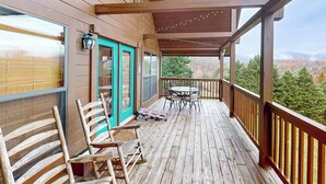 A wooden deck with a table and chairs, a covered area with a view of trees and mountains.