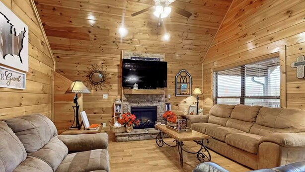 A cozy log cabin interior with a fireplace, two sofas, a television, and a ceiling fan.