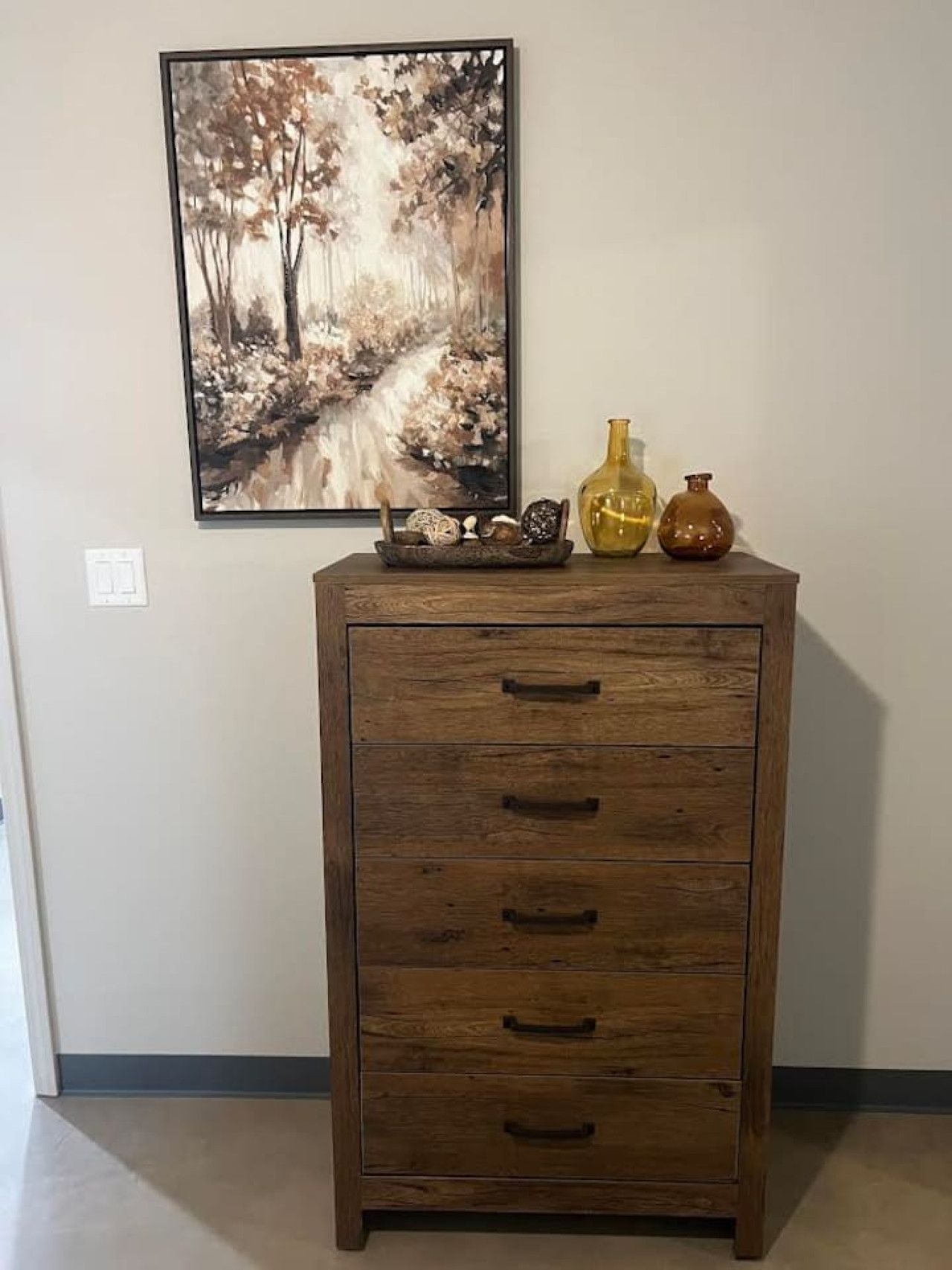 Un meuble en bois avec des tiroirs et une étagère, un tableau encadré représentant une scène de forêt, et deux vases décoratifs placés sur le dessus.