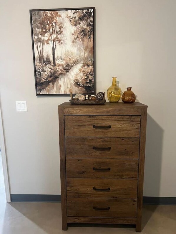 A wooden cabinet with drawers and a shelf, a framed painting of a forest scene, and two decorative vases on top.