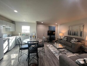 A modern kitchen with white cabinets, a dining area with a round table and chairs, a living room with a sofa and a TV, and a large painting on the wall.