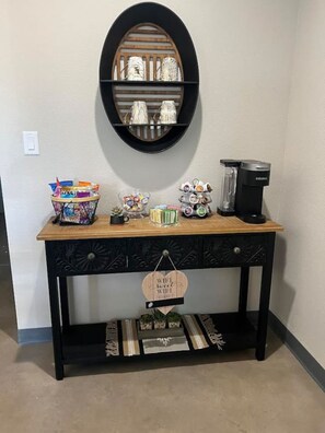 A black console table with a wooden top, a coffee maker, a basket of snacks, a small plant, and a decorative item hanging from the front.