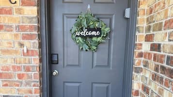 Una puerta gris con un cartel que dice 'welcome' y una corona de flores, ubicada en una pared de ladrillo.