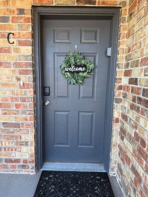 A grey door with a 'welcome' sign and a wreath, set in a brick wall. - Your Peaceful Retreat (Portales)