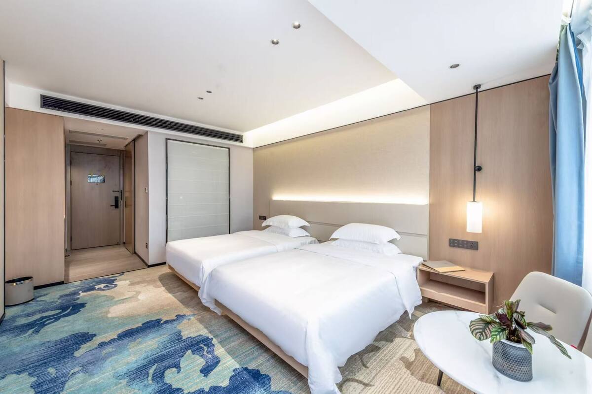 A modern hotel room with two beds, a small table, a chair, and a potted plant.