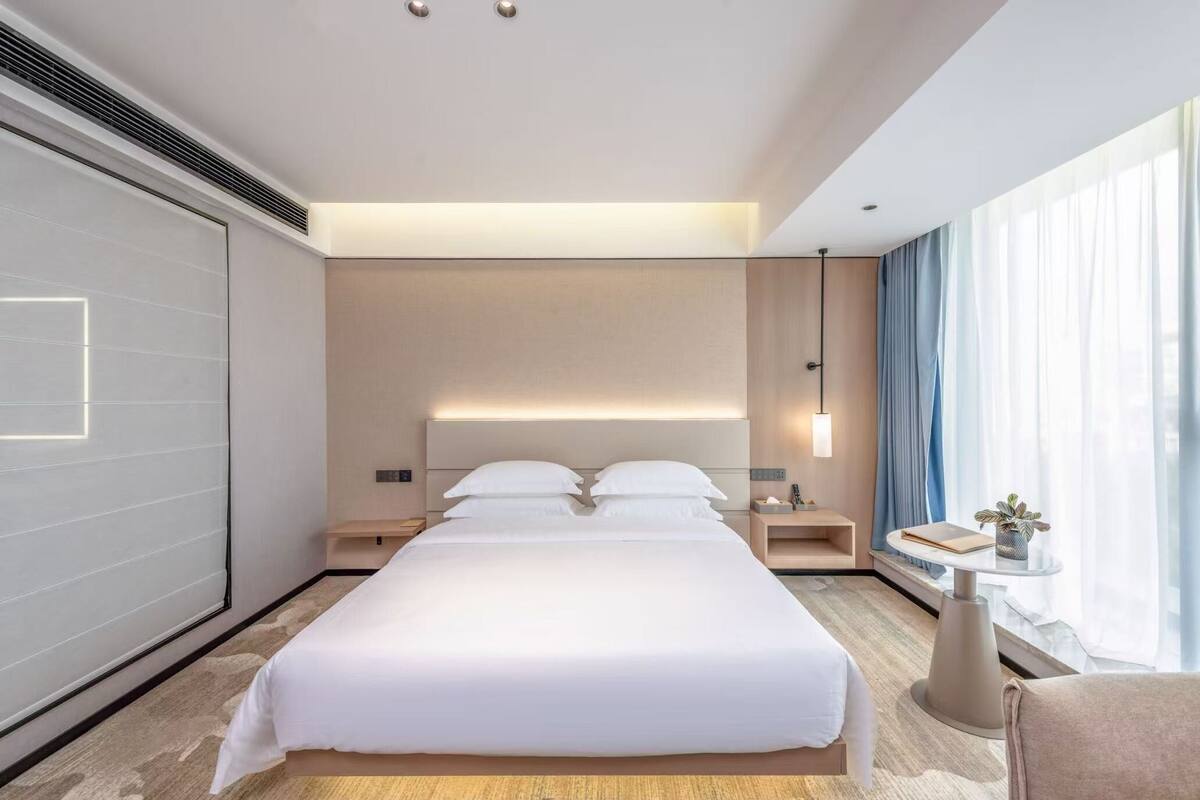 A modern hotel room with a large bed, bedside tables, a small table with a plant, and a large window with curtains.