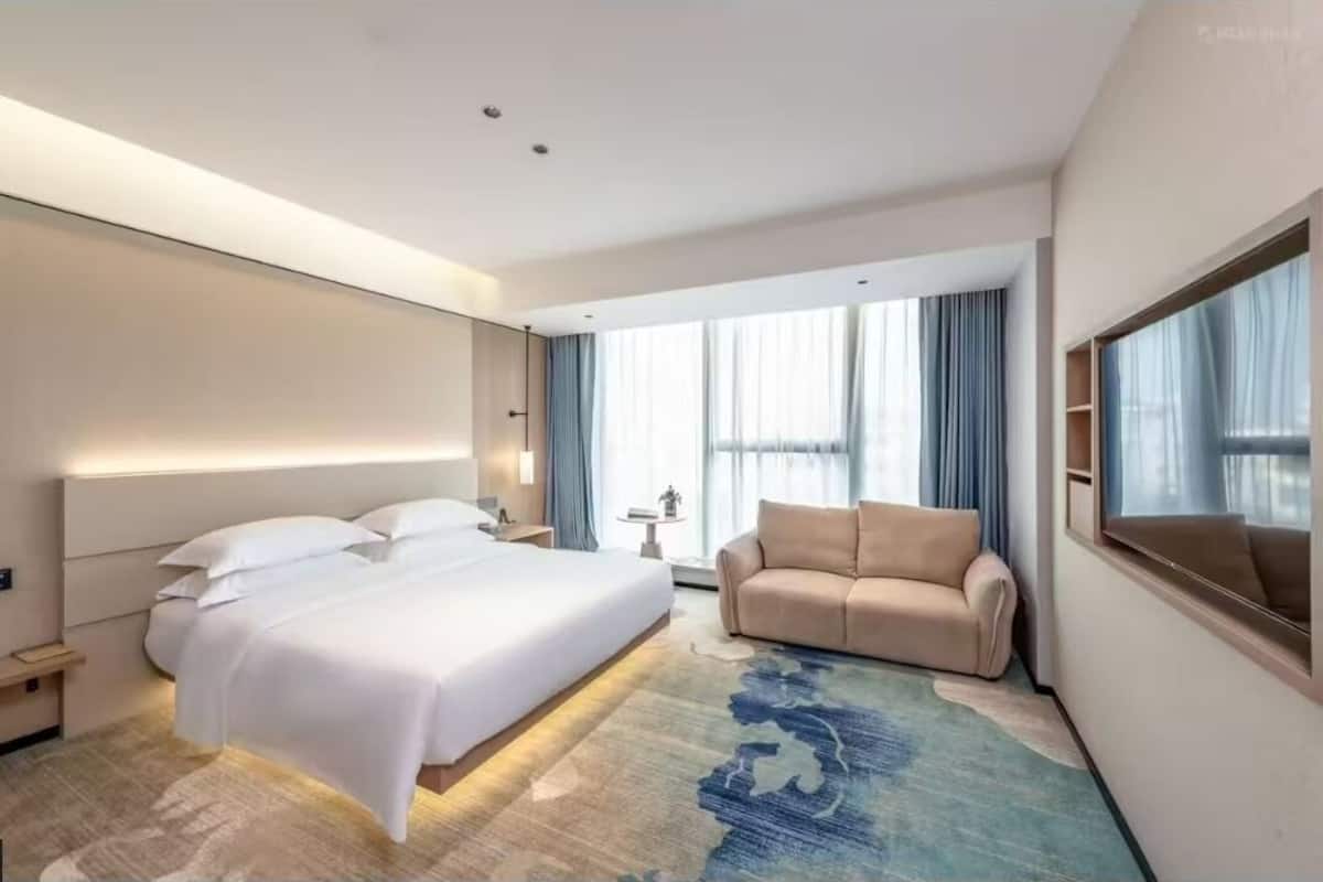 A modern hotel room with a large bed, a sofa, a small table, and a large window with curtains.