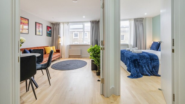 Una habitación de hotel moderna con sofá, una mesa de comedor, una cama con cubrecama azul y una planta en maceta.