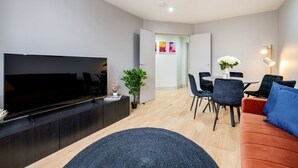Una sala moderna con un televisor de pantalla plana, un mueble de entretenimiento de madera oscura, una planta en maceta y un comedor con mesa y sillas.