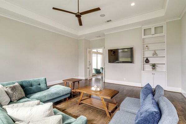 A spacious living room with a wooden coffee table, teal and gray sofas, a ceiling fan, and a TV mounted on the wall. - 9 Mi to Downtown Houston: Spacious Group Getaway (Houston)