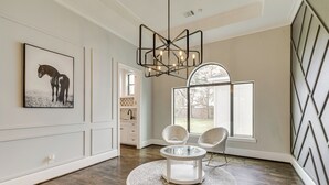 Un'ampia stanza con pavimento in parquet, un tavolo rotondo in vetro, due sedie bianche, un moderno lampadario e una grande finestra con vista sull'esterno.