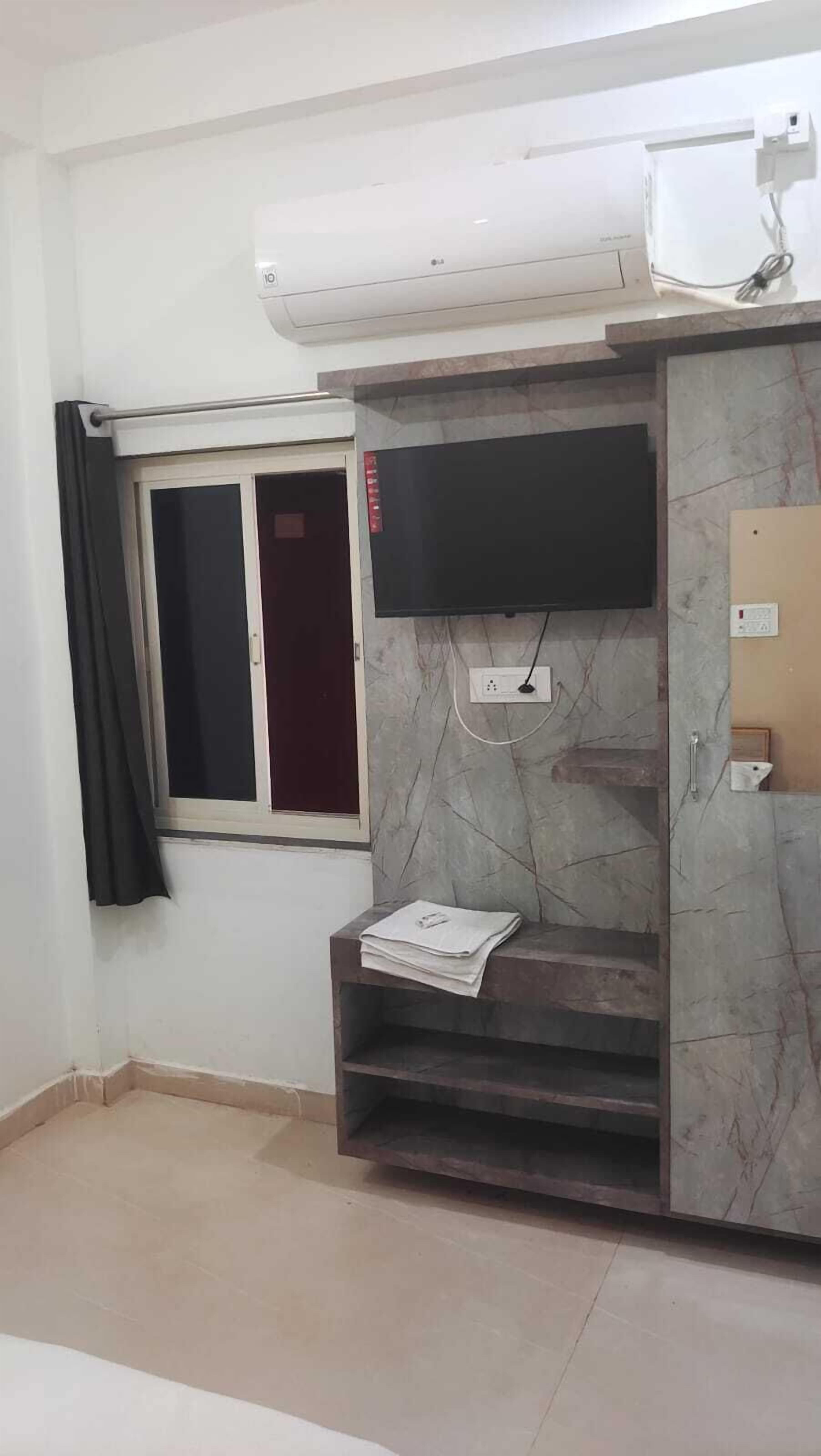 A room with a flat-screen TV mounted on a marble-patterned cabinet, a window with a curtain, and a wall-mounted air conditioner.