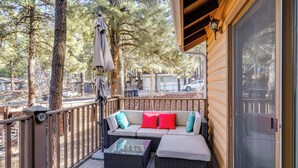 Une terrasse en bois avec un canapé et deux poufs, une table en verre et une rambarde en bois.