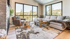 A modern living room with a fireplace, large windows, a sofa, and a coffee table. - Near Hiking & Biking: Winter Park Mtn-view Haven! (Winter Park)