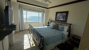 Un dormitorio con una cama, un ventilador de techo, una ventana con cortinas, un cuadro en la pared y una mesita.