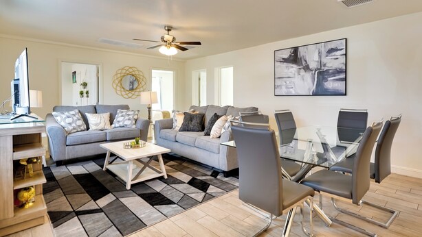 A modern living room with a grey sofa, a coffee table, a dining table with chairs, a ceiling fan, and artwork on the walls.