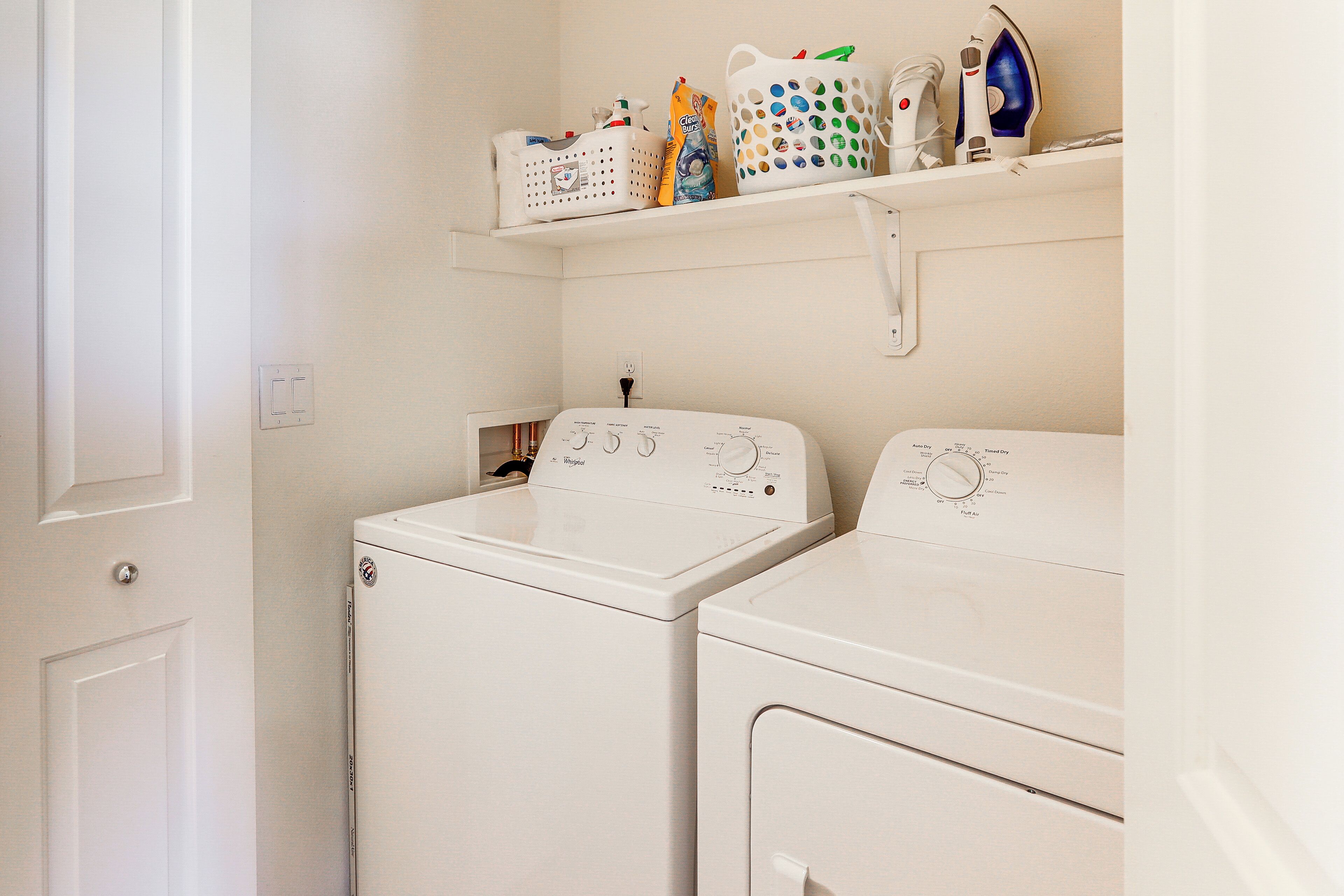 A laundry room with two white front-loading washing machines and a shelf above containing laundry supplies.