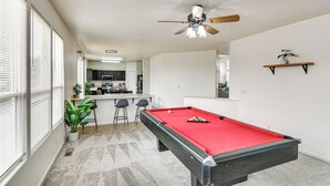 Een biljarttafel in een ruimte met een plafondventilator, een keuken op de achtergrond en een kamerplant.