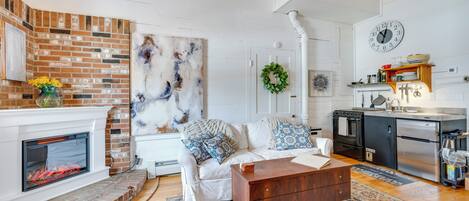 A living room with a fireplace, a white couch with blue and white pillows, a coffee table, and a kitchen area with a clock and appliances.