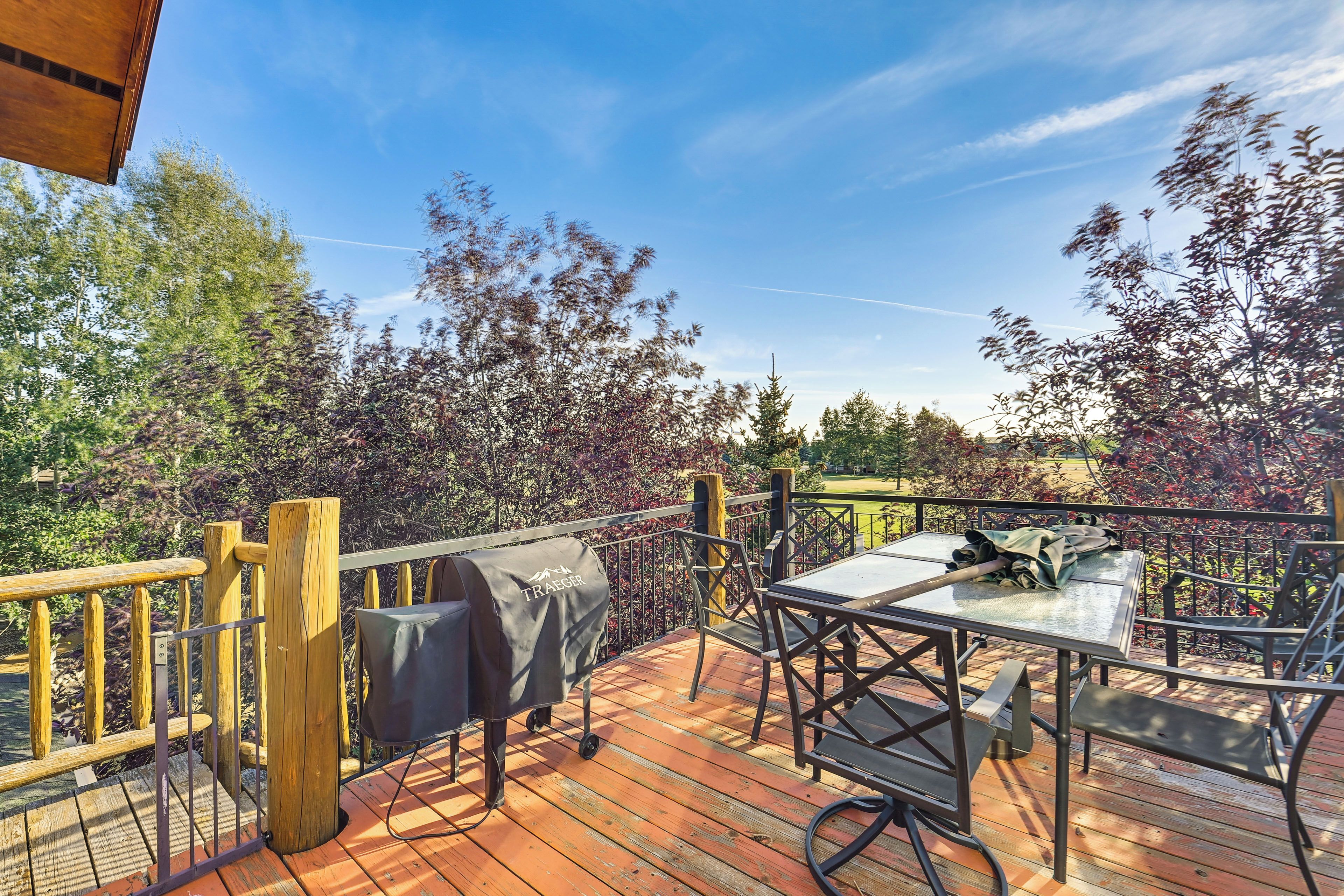 Una terraza al aire libre con una parrilla, una mesa y sillas, rodeada de árboles y cielo despejado.