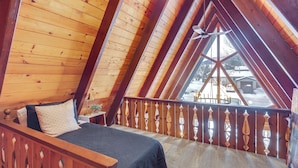 A bedroom with a wooden sloped ceiling, a bed with a dark blanket, and a view of a snowy landscape through a balcony railing. - Red Lodge Cabin w/ Hot Tub ~ 1/2 Mi to Downtown! (Red Lodge)