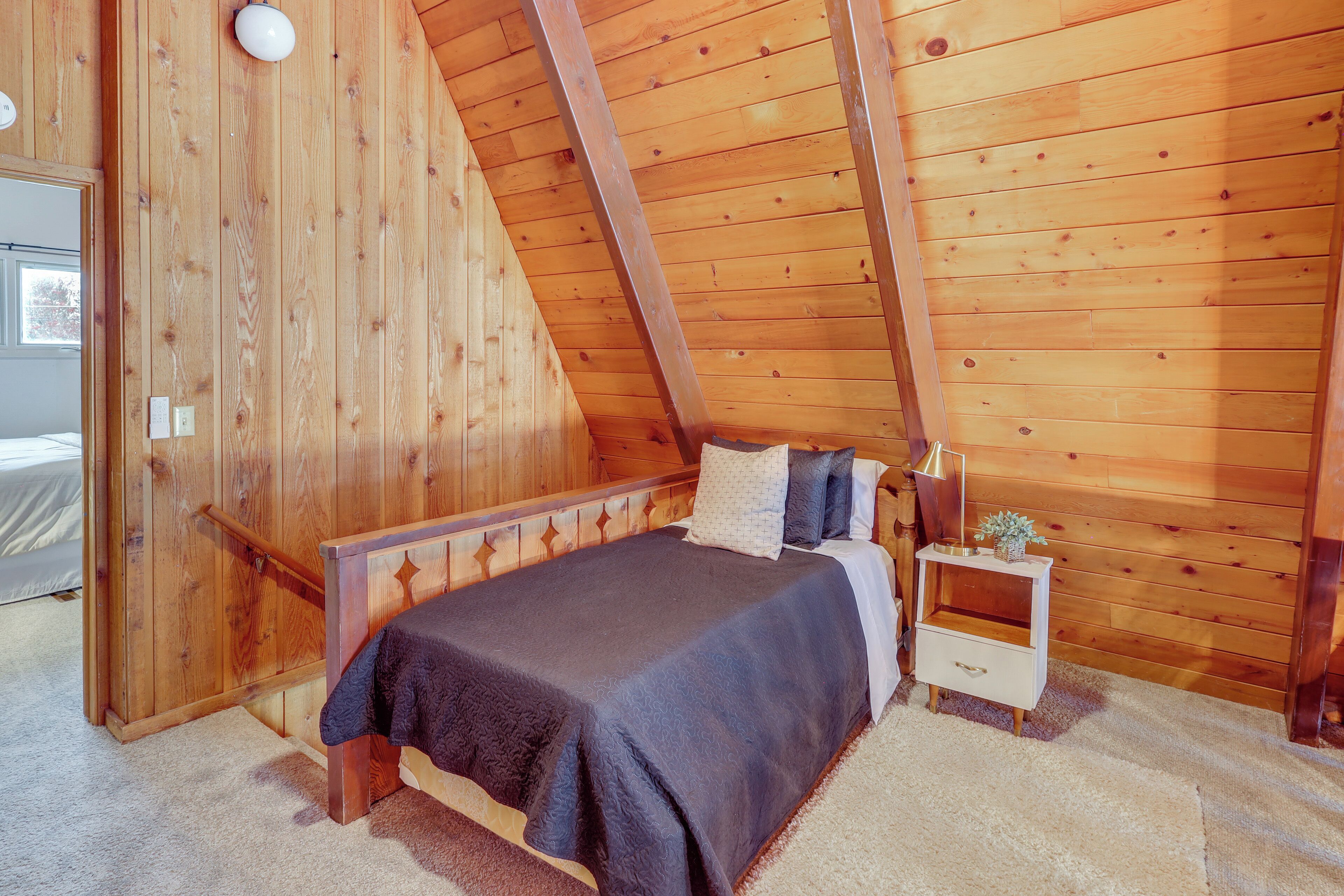 Ein Schlafzimmer mit holzverkleideter Schräge, einem Bett mit dunkler Bettdecke, einem Nachttisch mit Lampe und einer kleinen Pflanze.