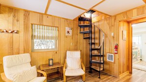 A wooden interior with a spiral staircase, two white cushioned chairs, a small table, and a window with blinds.
