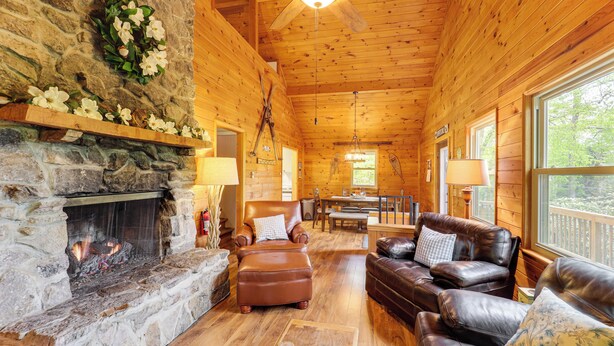 A cozy cabin interior with a stone fireplace, leather furniture, and large windows offering a view of the outdoors.