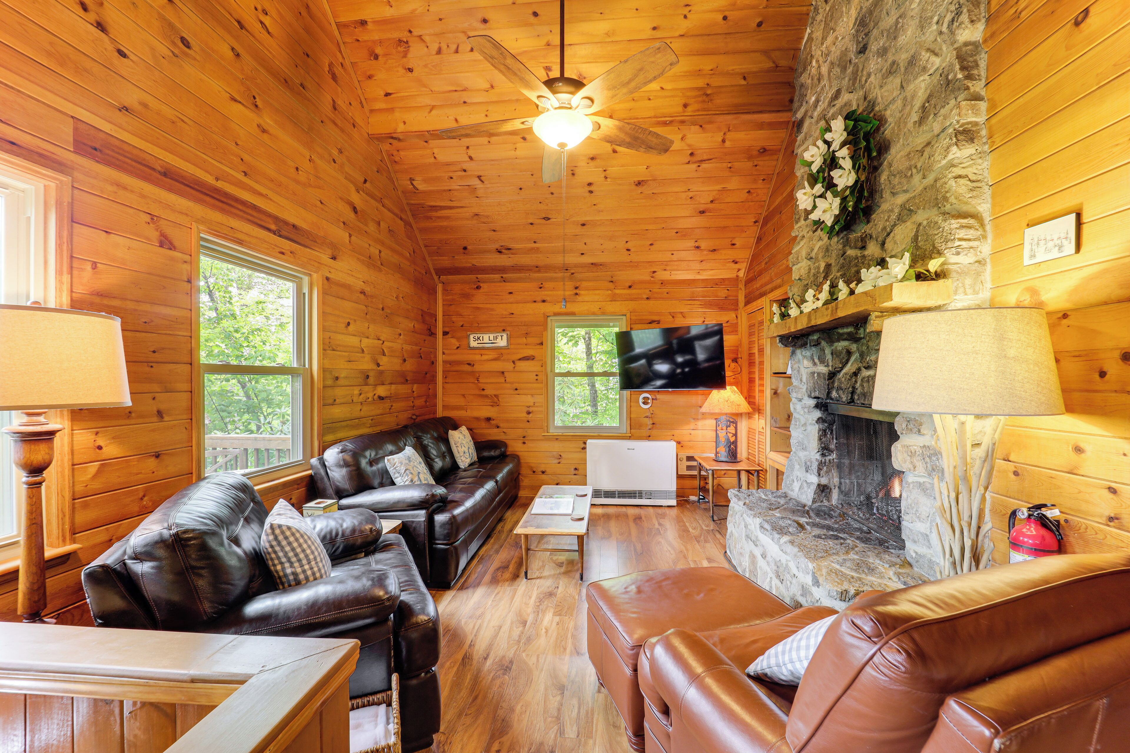 A cozy cabin interior with wooden walls, a stone fireplace, leather sofas, and a ceiling fan.