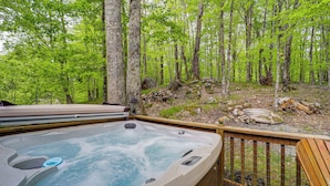 A hot tub surrounded by a wooden deck, set in a forested area with trees and a stone fire pit.