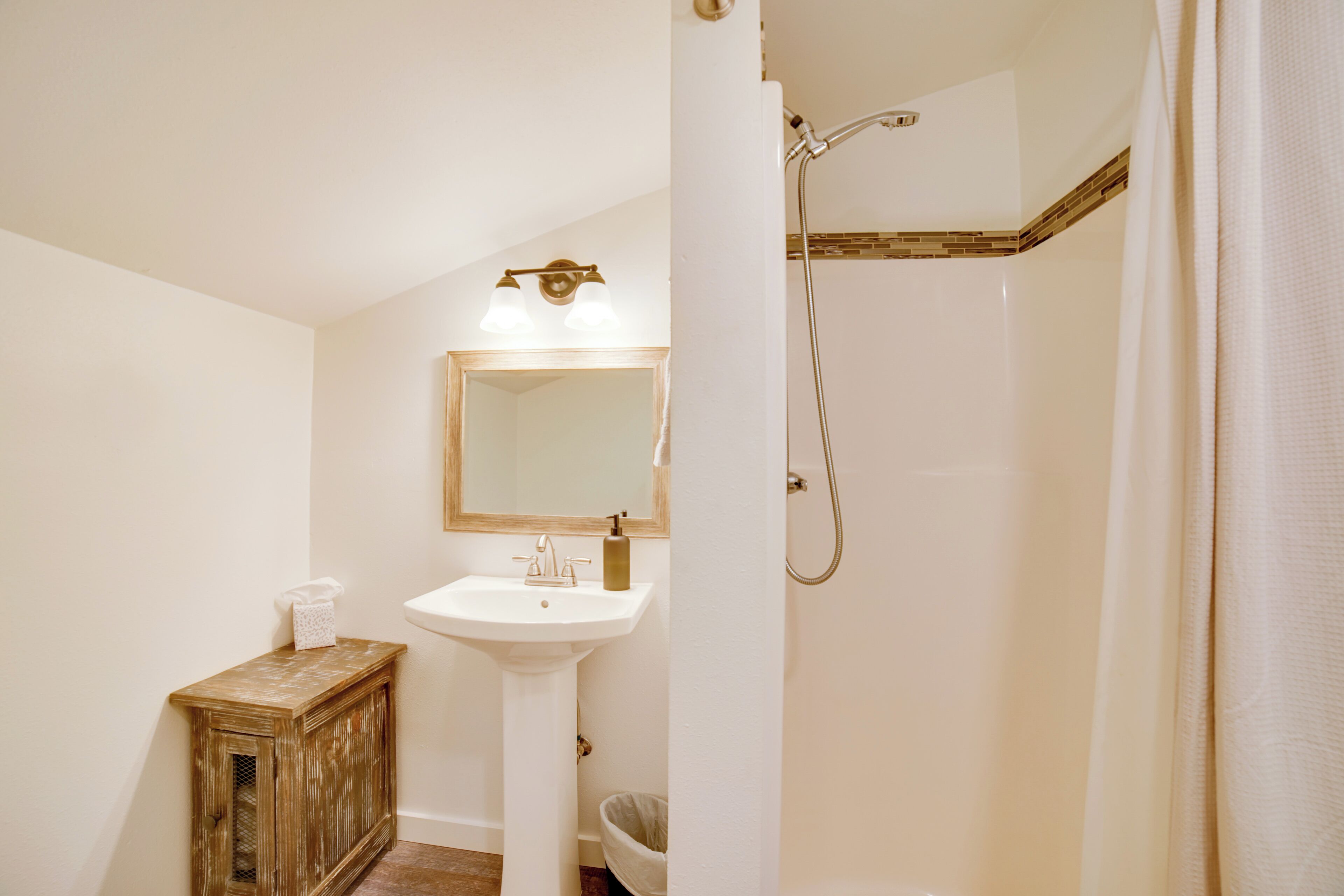 Une salle de bain avec un lavabo sur colonne, un miroir, une douche et un petit meuble.