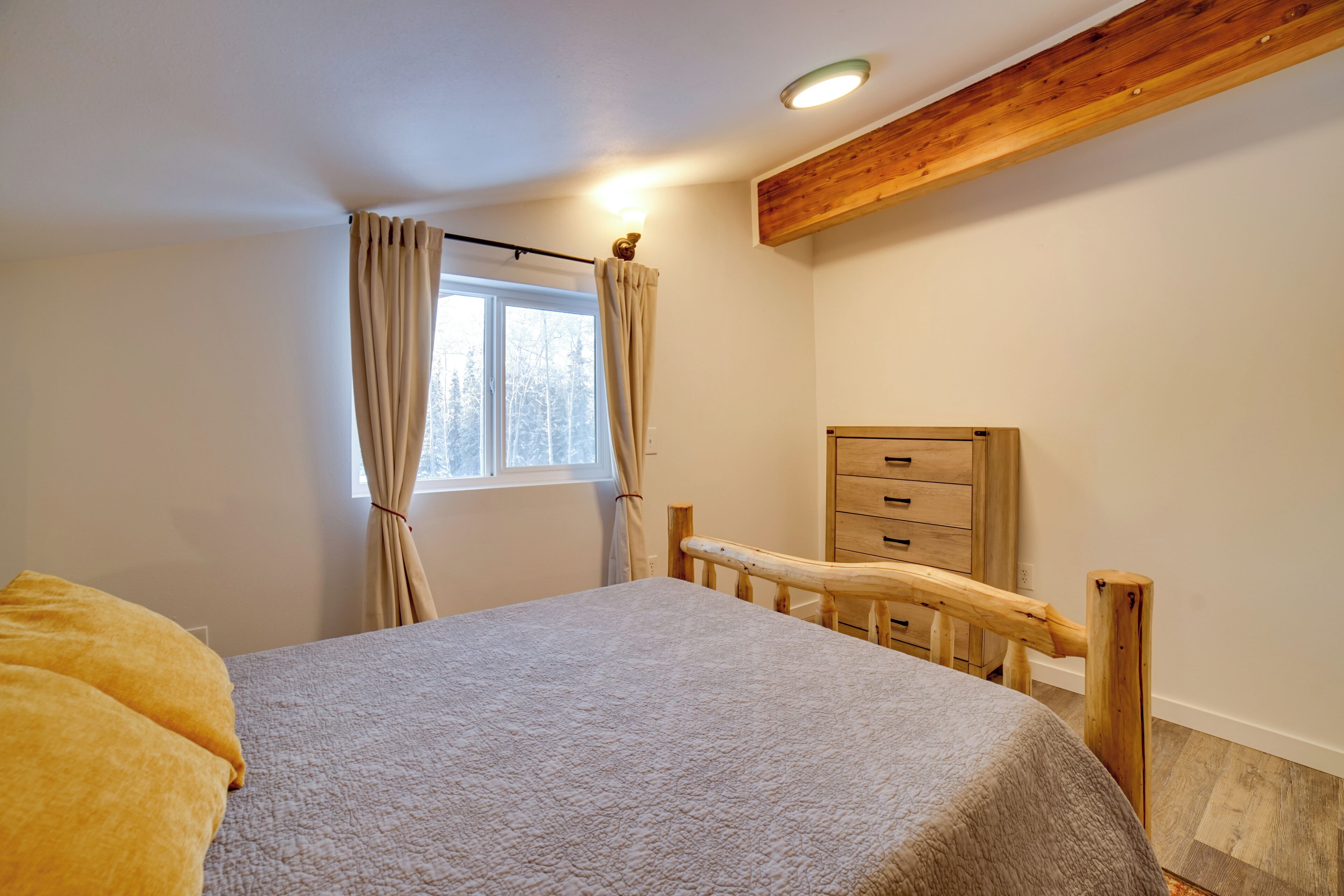 A bedroom with a bed, wooden headboard, a window with curtains, a wooden dresser, and a ceiling light.