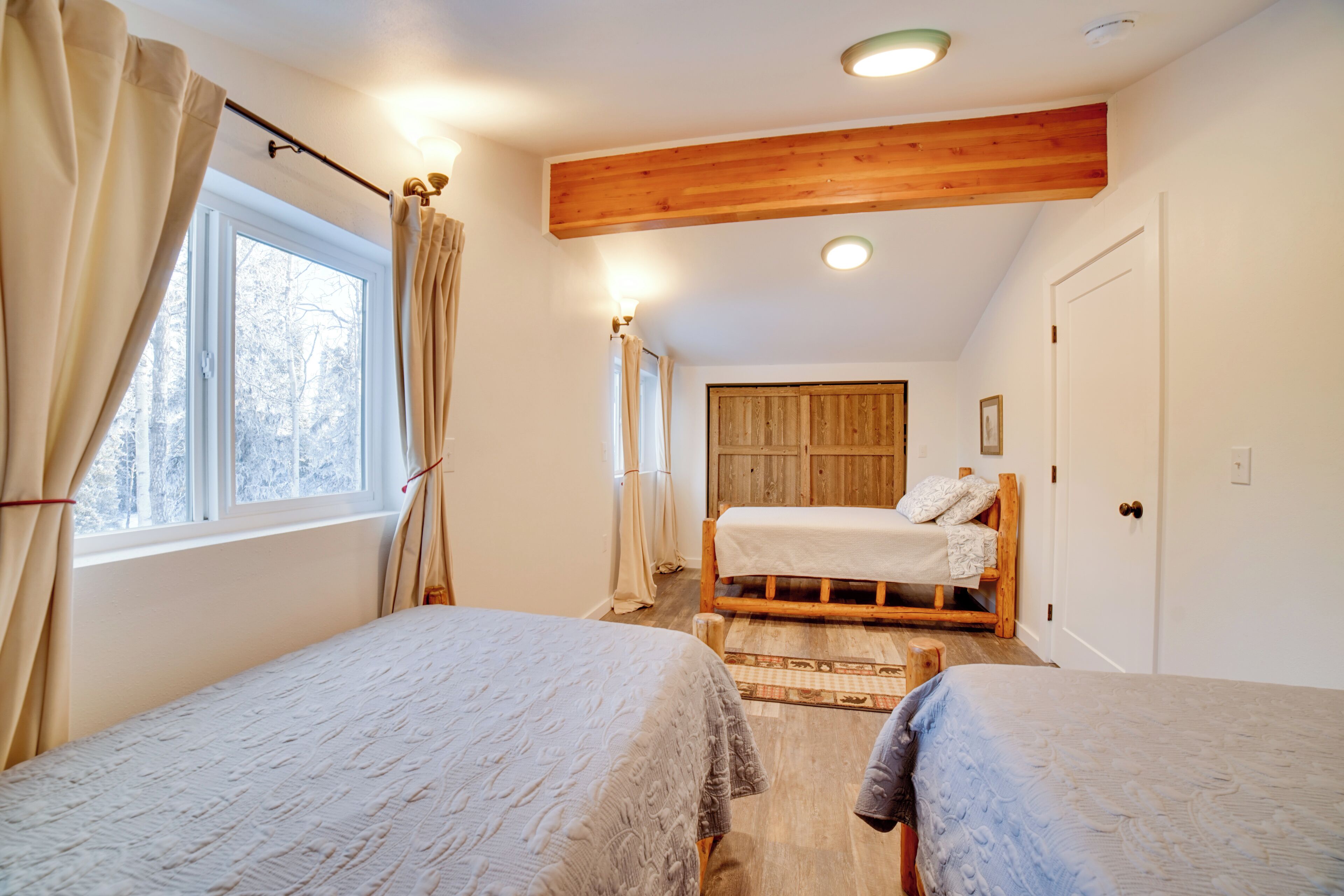 A bedroom with two beds, a wooden ceiling, a window with curtains, and a door.