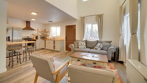 Een ruime woonkamer met een bank, salontafel en barstoelen. In de achtergrond is een keuken met moderne apparatuur zichtbaar.