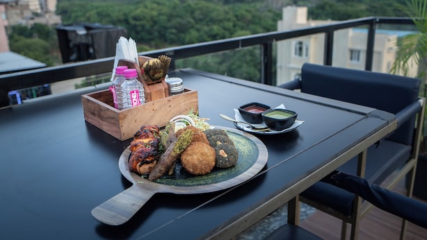 A dining table with a variety of food items, including grilled meat and fried snacks, on a plate. There is a bottle of water and condiments on the table. The setting is on a balcony with a view of greenery and buildings in the background. - THE COURTYARD - Beyond Bar & Club (Udaipur)