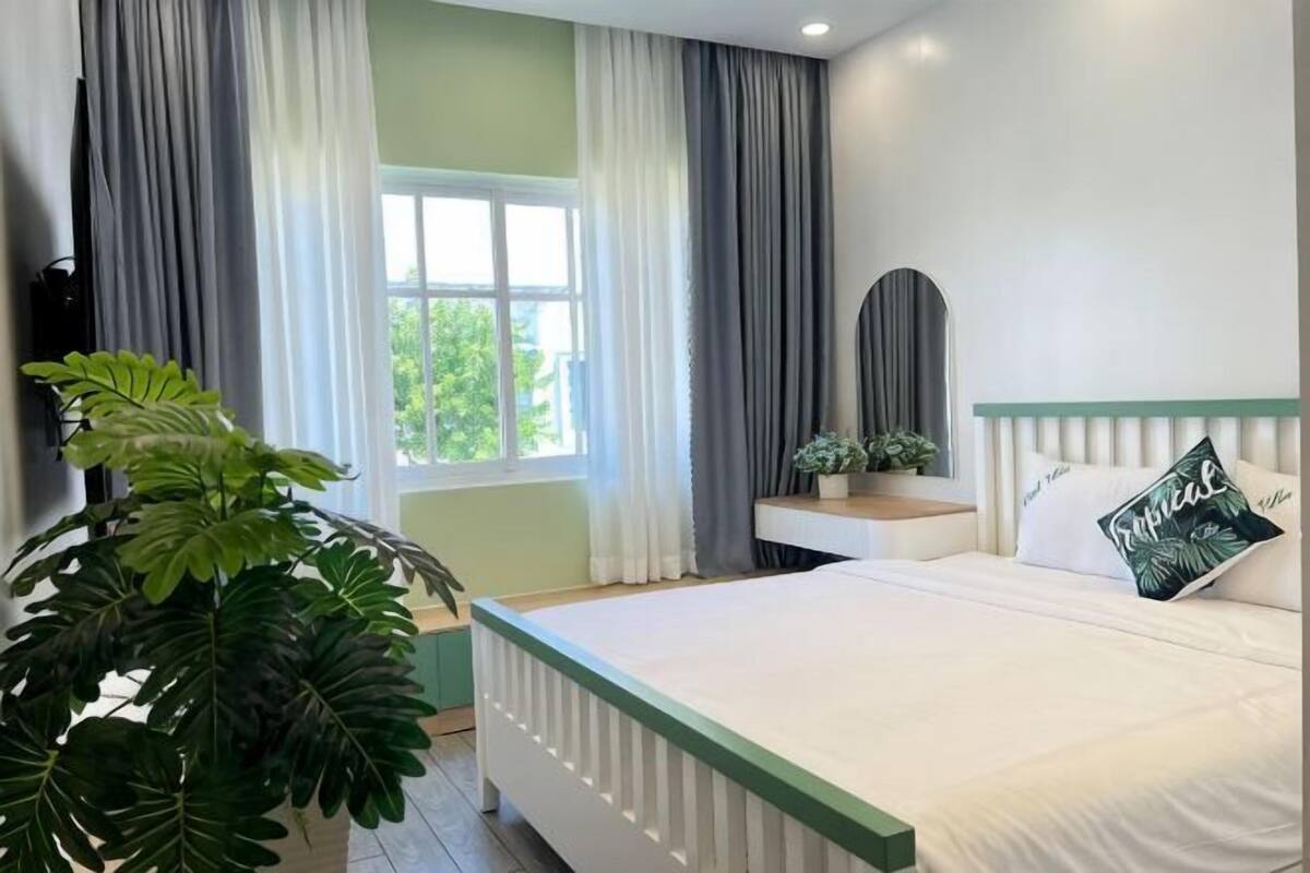 A bedroom with a bed, a bedside table, a mirror, a window with curtains, and a potted plant.
